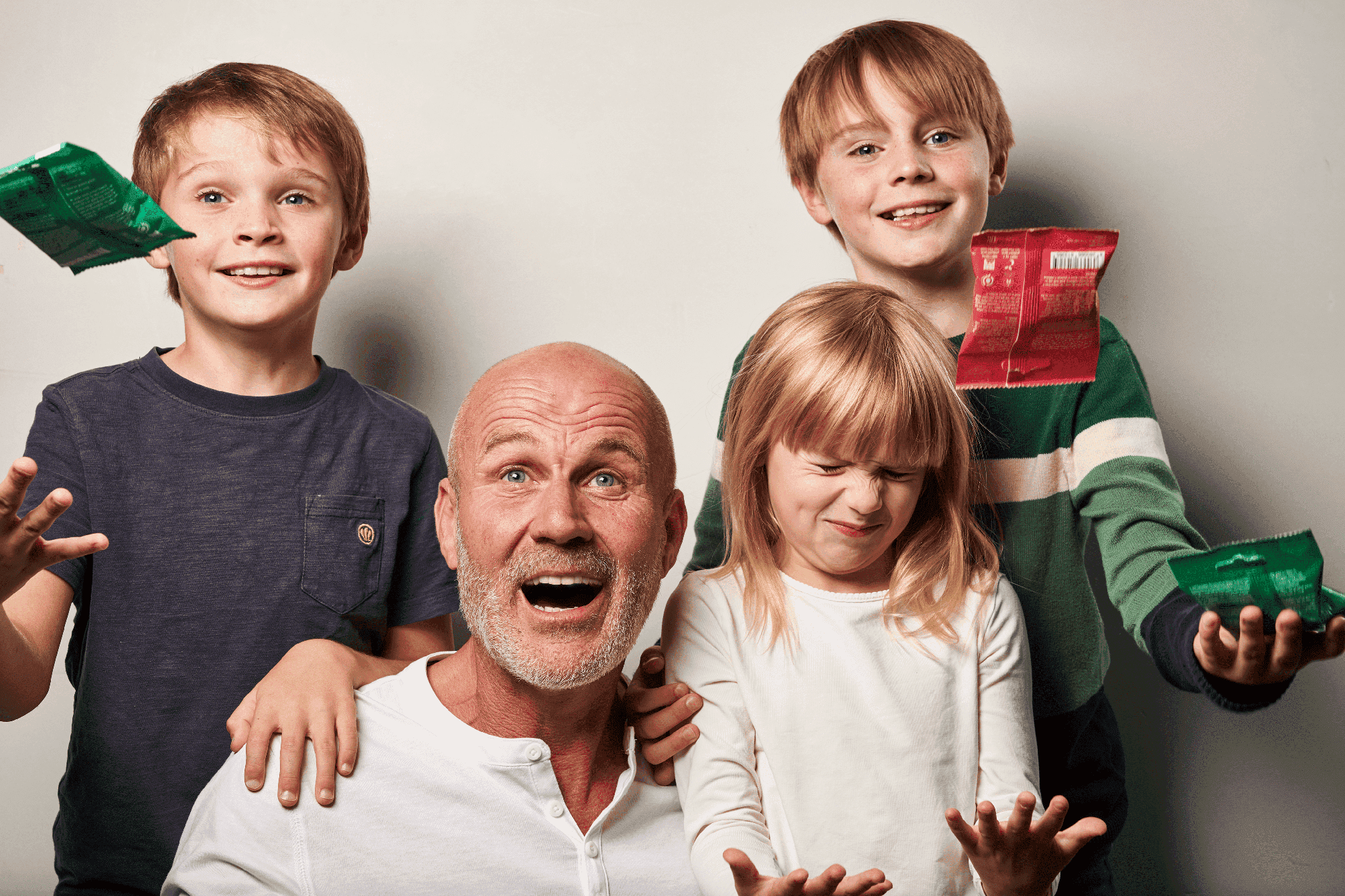 Ein lächelnder älterer Mann und drei fröhliche Kinder, zwei Jungen und ein Mädchen, werfen Bonbonpapier in die Luft - der perfekte Spaß für Familien, die Denttabs Zahnputztabletten als Zahnpasta-Alternative entdecken.