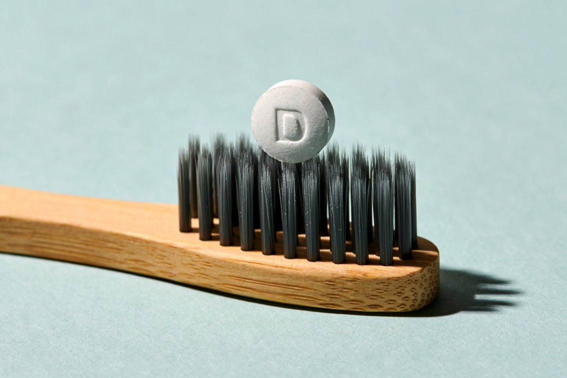 Eine runde weiße Denttabs Zahnputztablette mit einem "D" ruht auf den Borsten einer Bambuszahnbürste vor einem blassgrünen Hintergrund - eine trockene, vegane Zahnpasta-Alternative.