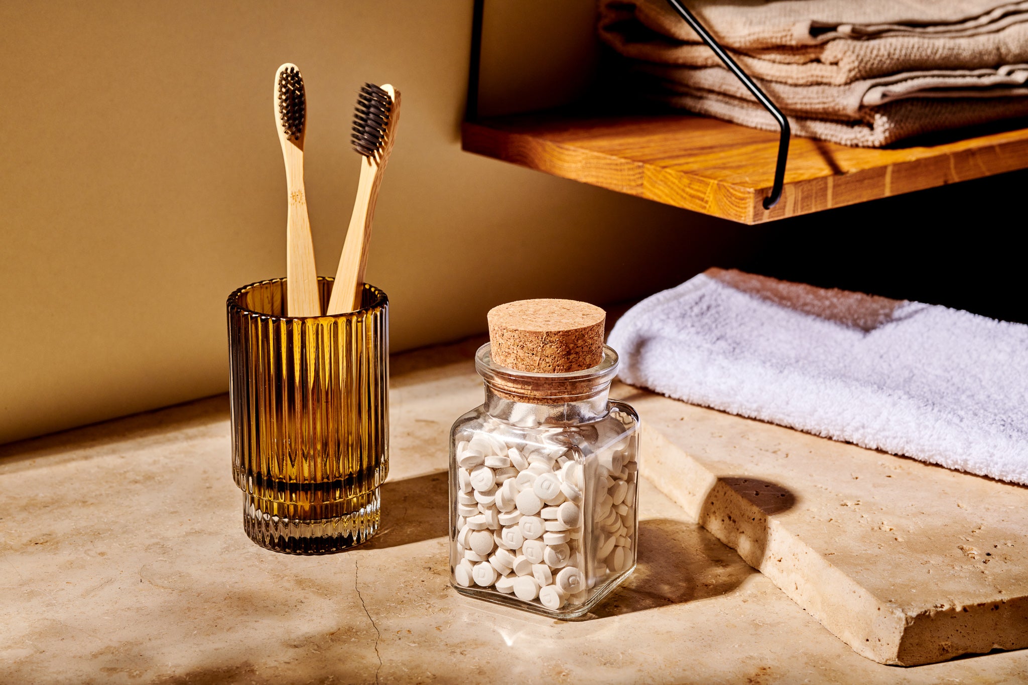 Zwei Bambuszahnbürsten in einem Glasbecher stehen neben einer Dose Denttabs Zahnputztabletten - einer idealen Zahnpasta-Alternative - auf einem Badezimmertisch, darüber liegen gefaltete Handtücher auf einem Holzregal.
