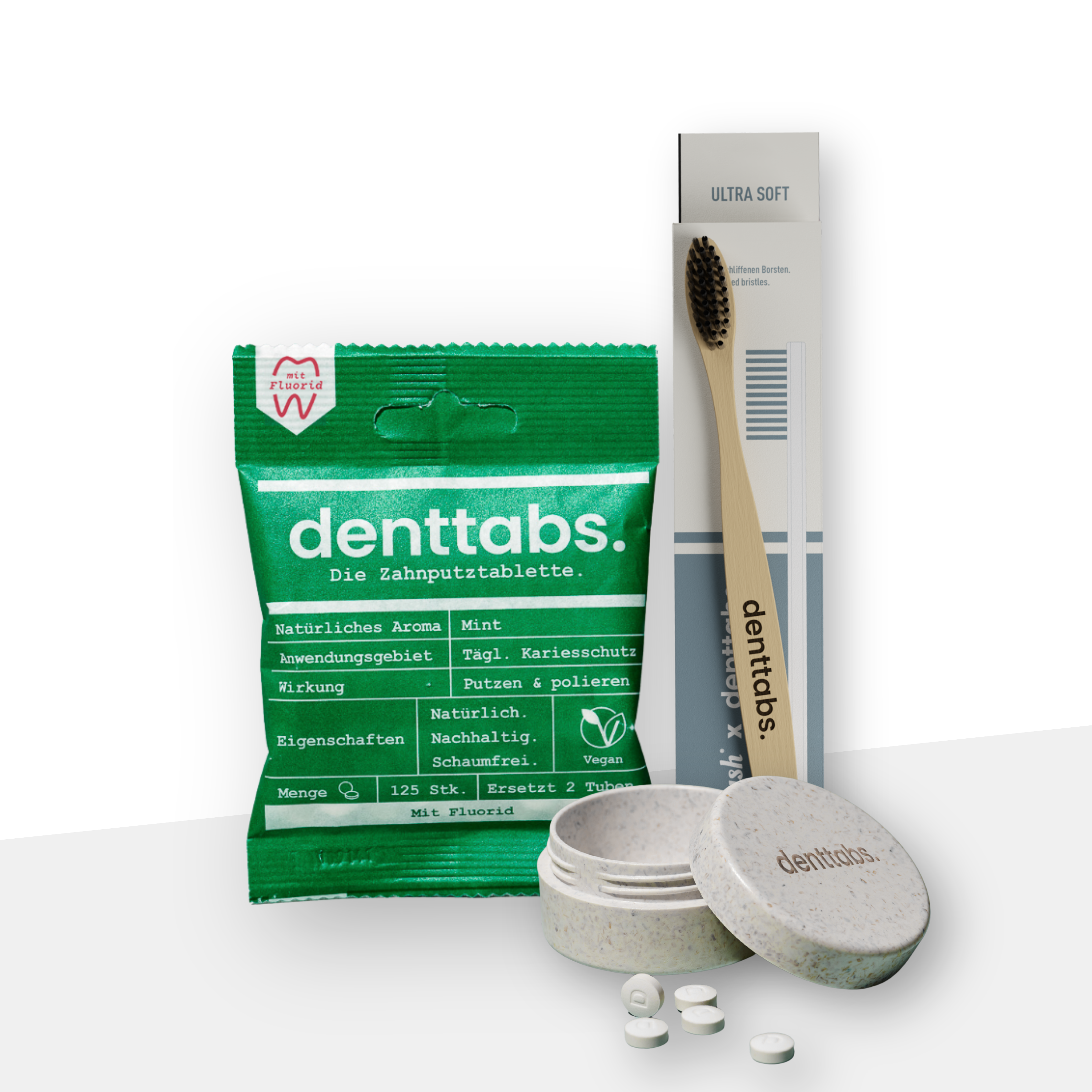 Auf dem Bild ist das Reiseset von Dentabs vor einem hellen Hintergrund abgebildet: Eine grüne Tüte Denttabs, eine Bambuszahnbürste mit Verpackung und die Reisedose von Denttabs in grau / beige.