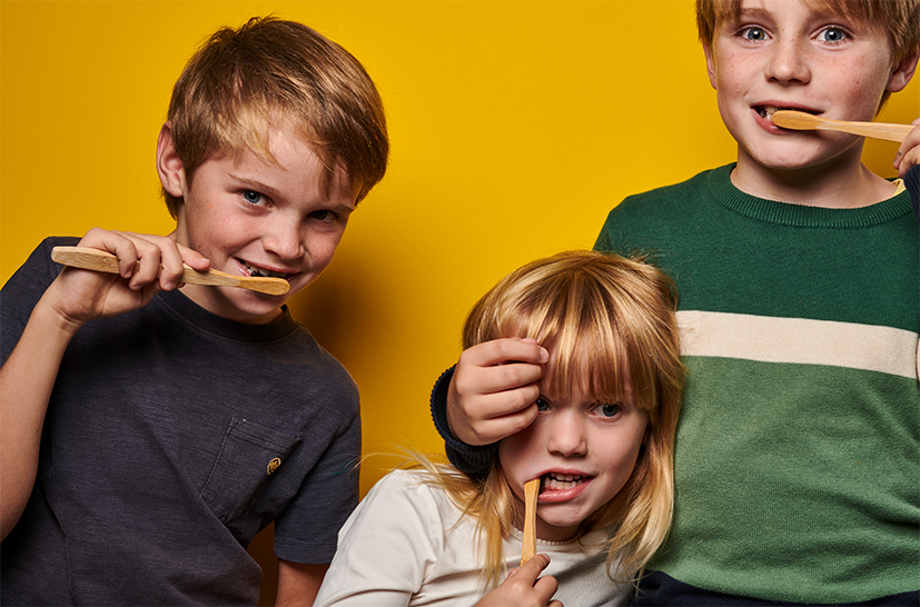 Drei Kinder lächeln in die Kamera, während sie sich mit Holzzahnbürsten vor einem gelben Hintergrund die Zähne putzen - ein idealer Moment, um Denttabs Zahnputztabletten als lustige, umweltfreundliche Alternative zu herkömmlicher Zahnpasta auszuprobieren.